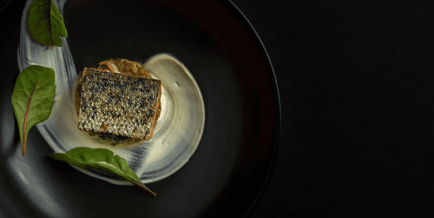 Plated gourmet fish dish with sauce and leafy greens on a dark background.