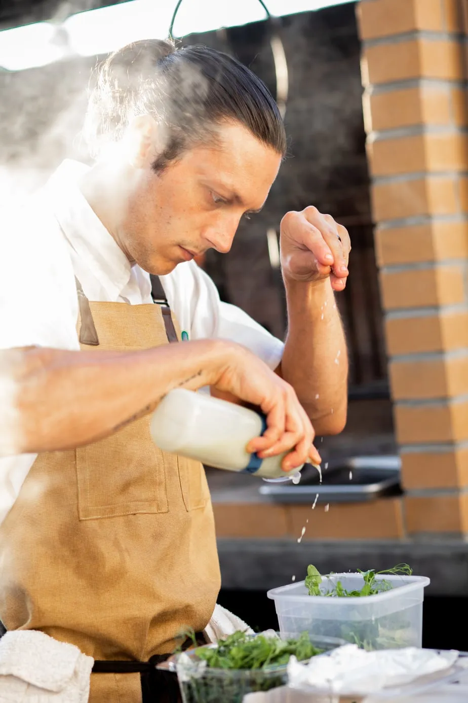 Chef in a brown apron carefully seasoning food with herbs and salt in a steamy kitchen.