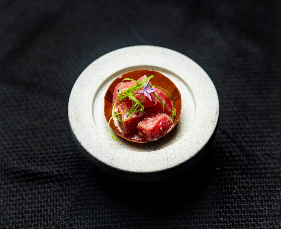 Gourmet dish featuring diced meat garnished with green herbs and a small purple flower on a round white plate.