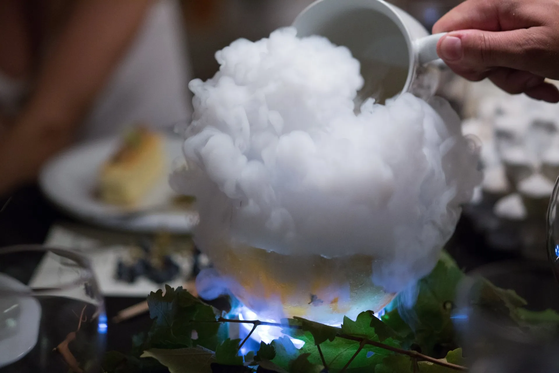 Hand pouring liquid to create a cloud of smoke or vapor over a bowl with decorative leaves and lights