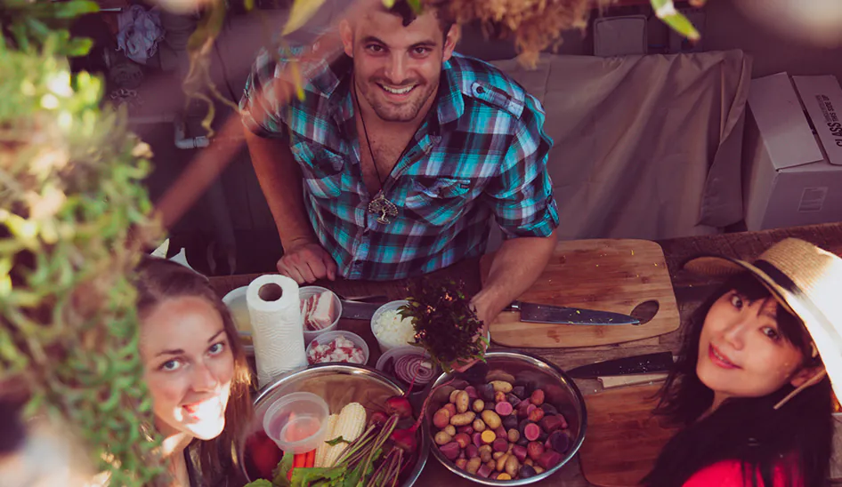 Three people smiling while preparing fresh vegetables and food outdoors.